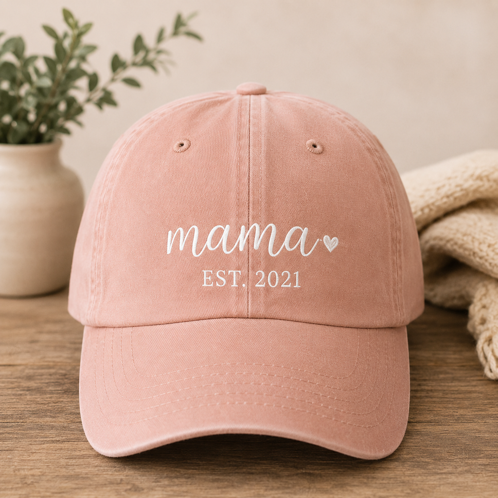 Pink cap with 'mama EST. 2021' text on a wooden surface with a plant and blanket in the background