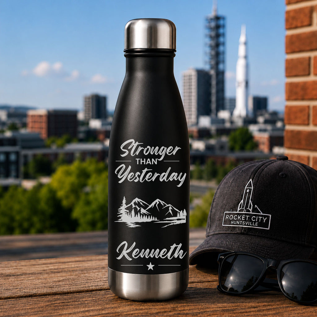 Black water bottle with 'Stronger than Yesterday' text and mountains, next to a black cap and sunglasses with a cityscape in the background.