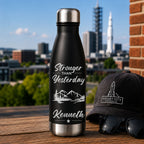 Black water bottle with 'Stronger than Yesterday' text and mountains, next to a black cap and sunglasses with a cityscape in the background.