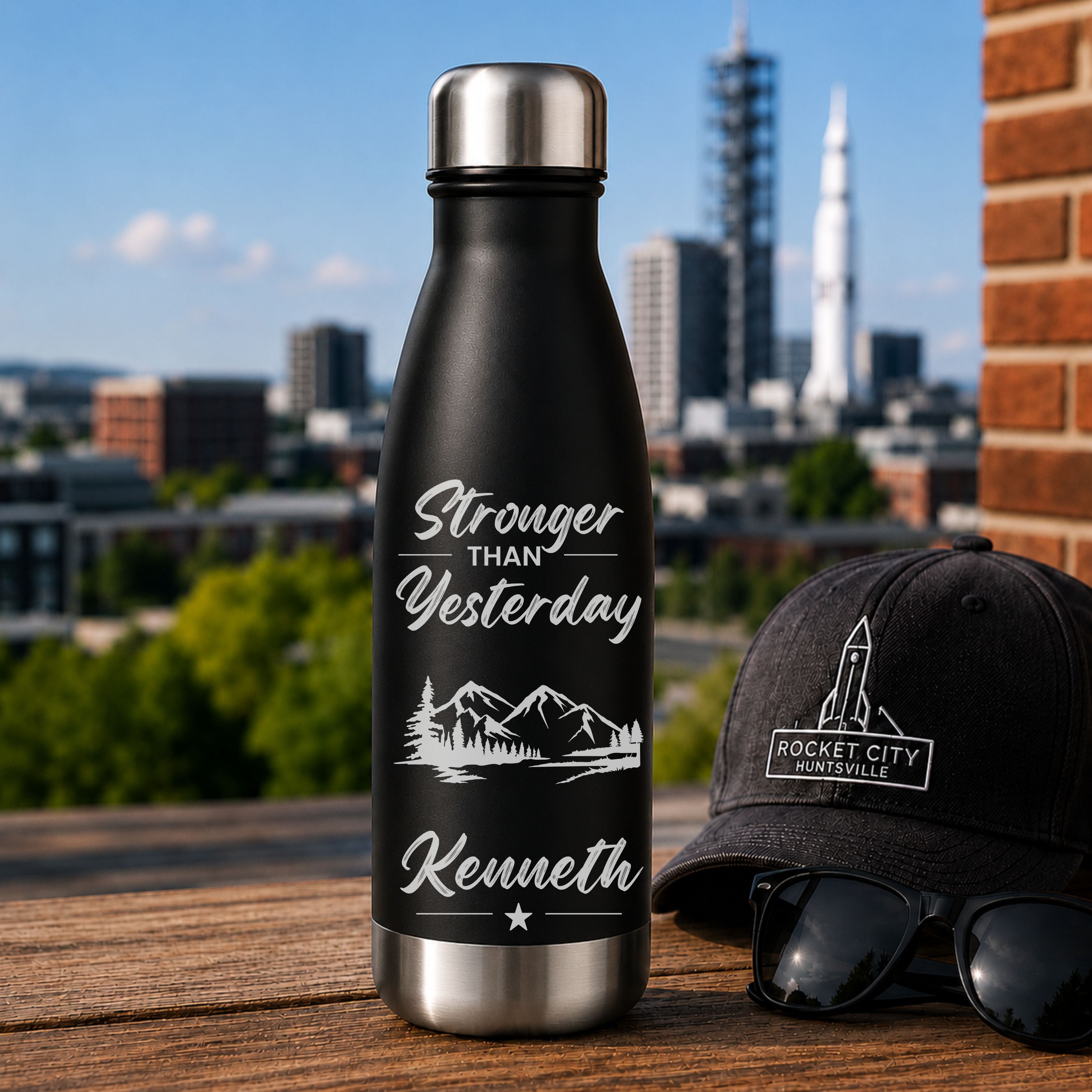 Black water bottle with 'Stronger than Yesterday' text and mountains, next to a black cap and sunglasses with a cityscape in the background.