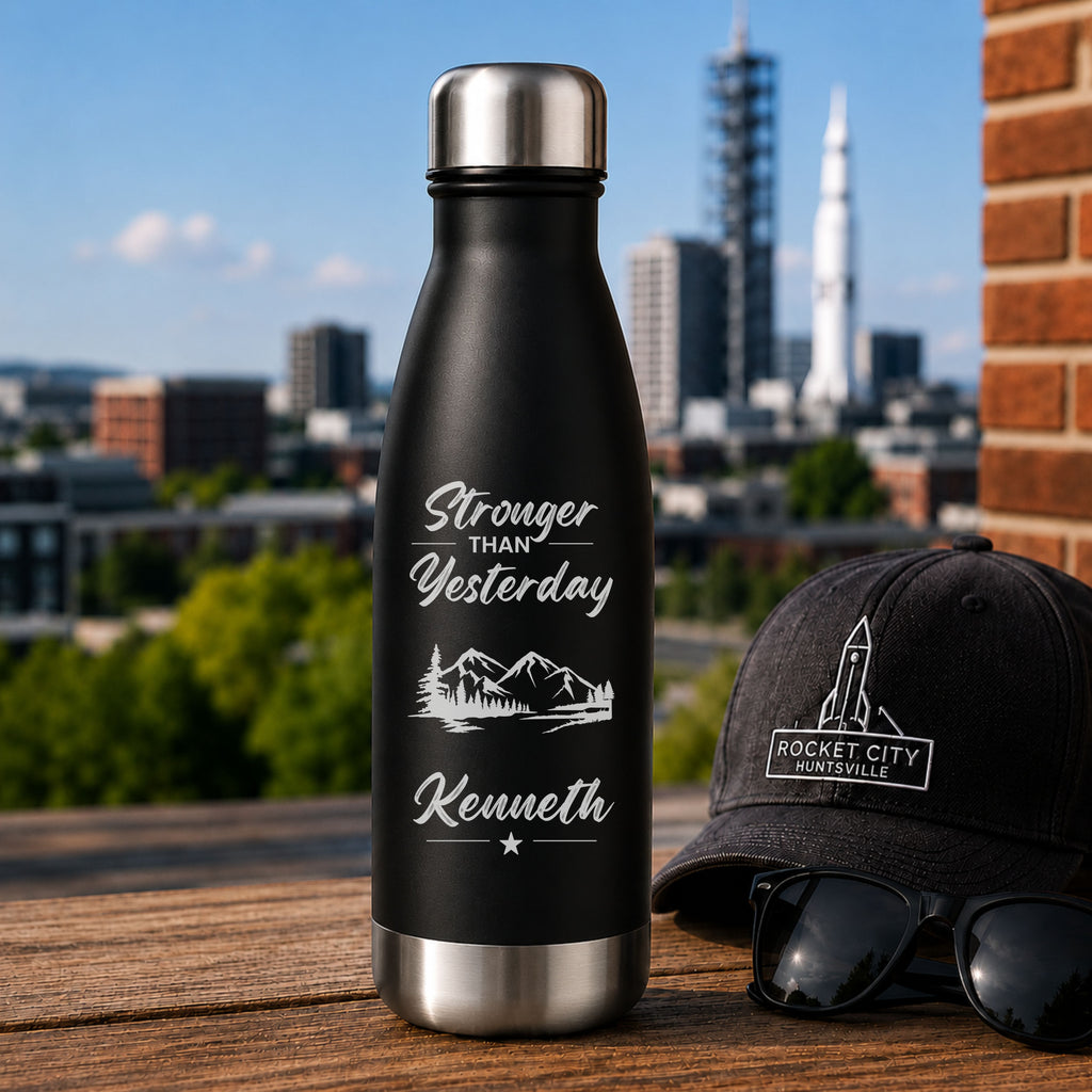 Black water bottle with 'Stronger than Yesterday' text and mountains, next to a black cap and sunglasses with a cityscape in the background.