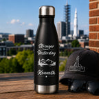 Black water bottle with 'Stronger than Yesterday' text and mountains, next to a black cap and sunglasses with a cityscape in the background.