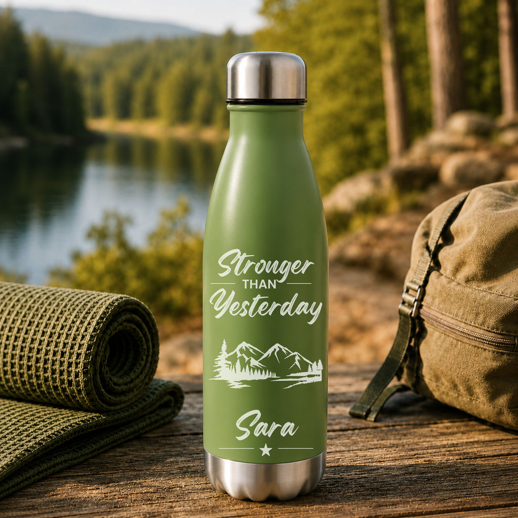 Green water bottle with 'Stronger than Yesterday' text and mountains placed outdoors with nature background.  