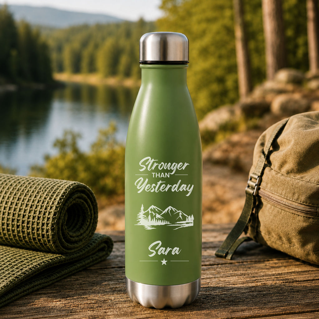 Green insulated bottle with 'Stronger Than Yesterday' text and mountain design, placed outdoors with a lake and forest in the background.