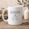 White mug with 'GRANDMA Loved By Sam Becca Julia John' text on a wooden surface with a neutral background