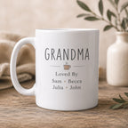 White mug with 'GRANDMA Loved By Sam Becca Julia John' text on a wooden surface with a neutral background