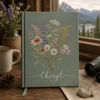 Green journal with floral design and 'Cheryl' on a wooden surface with a mountain view.