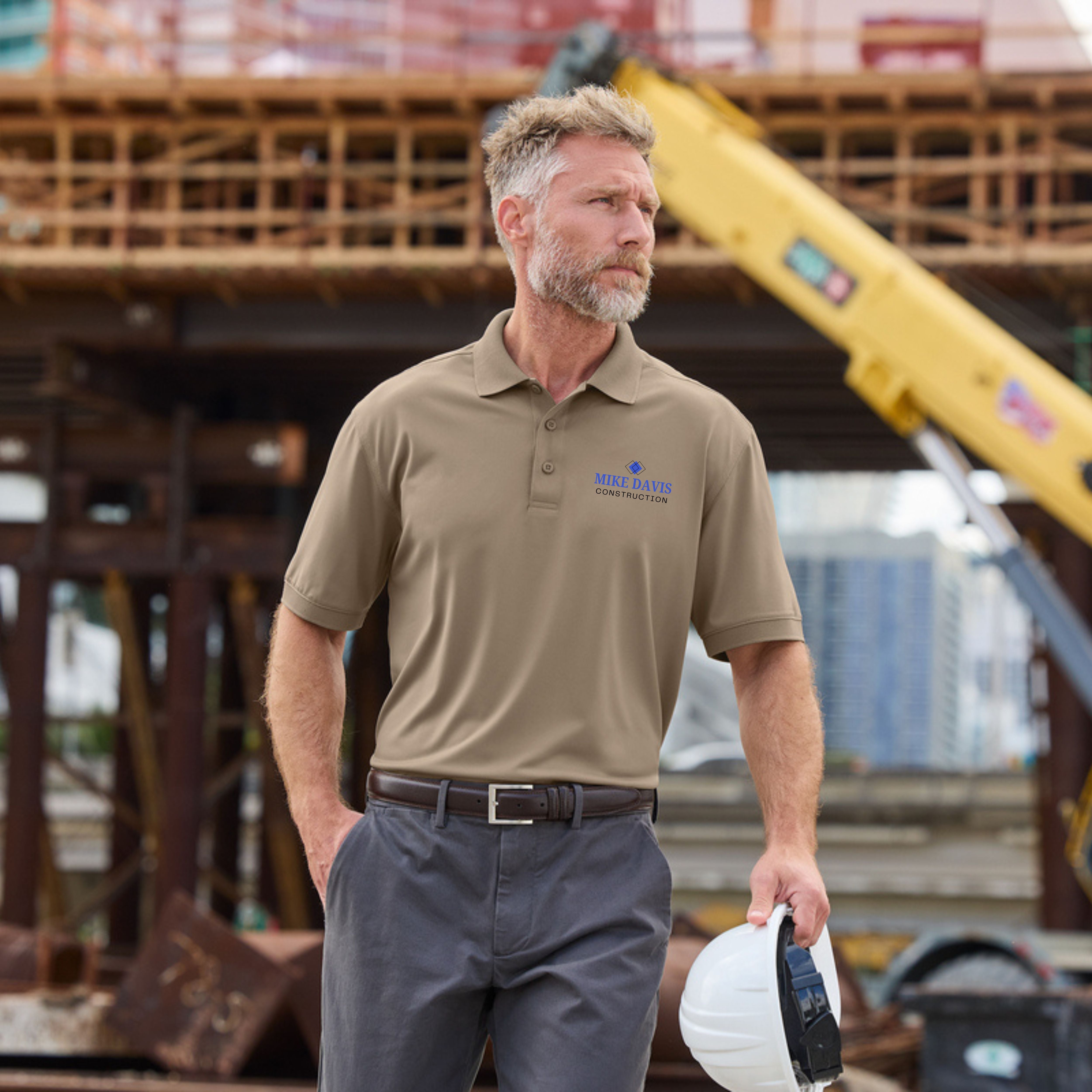 Man in a brown polo shirt and gray pants holding a hard hat on a construction site.