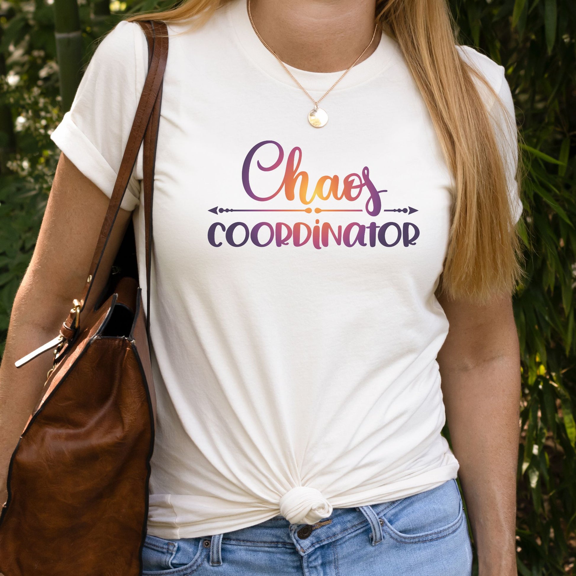 Woman standing with purse over shoulder wearing a nautral color t-shirt with Chaos Coordinator written in multicolored letters in center of shirt.