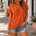 Woman wearing an orange t-shirt with text  "Running on Coffee and Grace" with kids names and denim shorts outdoors.