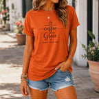 Woman wearing an orange t-shirt with text  "Running on Coffee and Grace" with kids names and denim shorts outdoors.