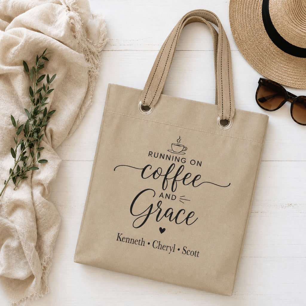 Beige tote bag with 'Running on Coffee and Grace' text and kids names, placed on a light wooden surface with a hat, sunglasses, and plant.