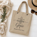 Beige tote bag with 'Running on Coffee and Grace' text and kids names, placed on a light wooden surface with a hat, sunglasses, and plant.