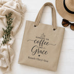 Beige tote bag with 'Running on Coffee and Grace' text and kids names, placed on a light wooden surface with a hat, sunglasses, and plant.