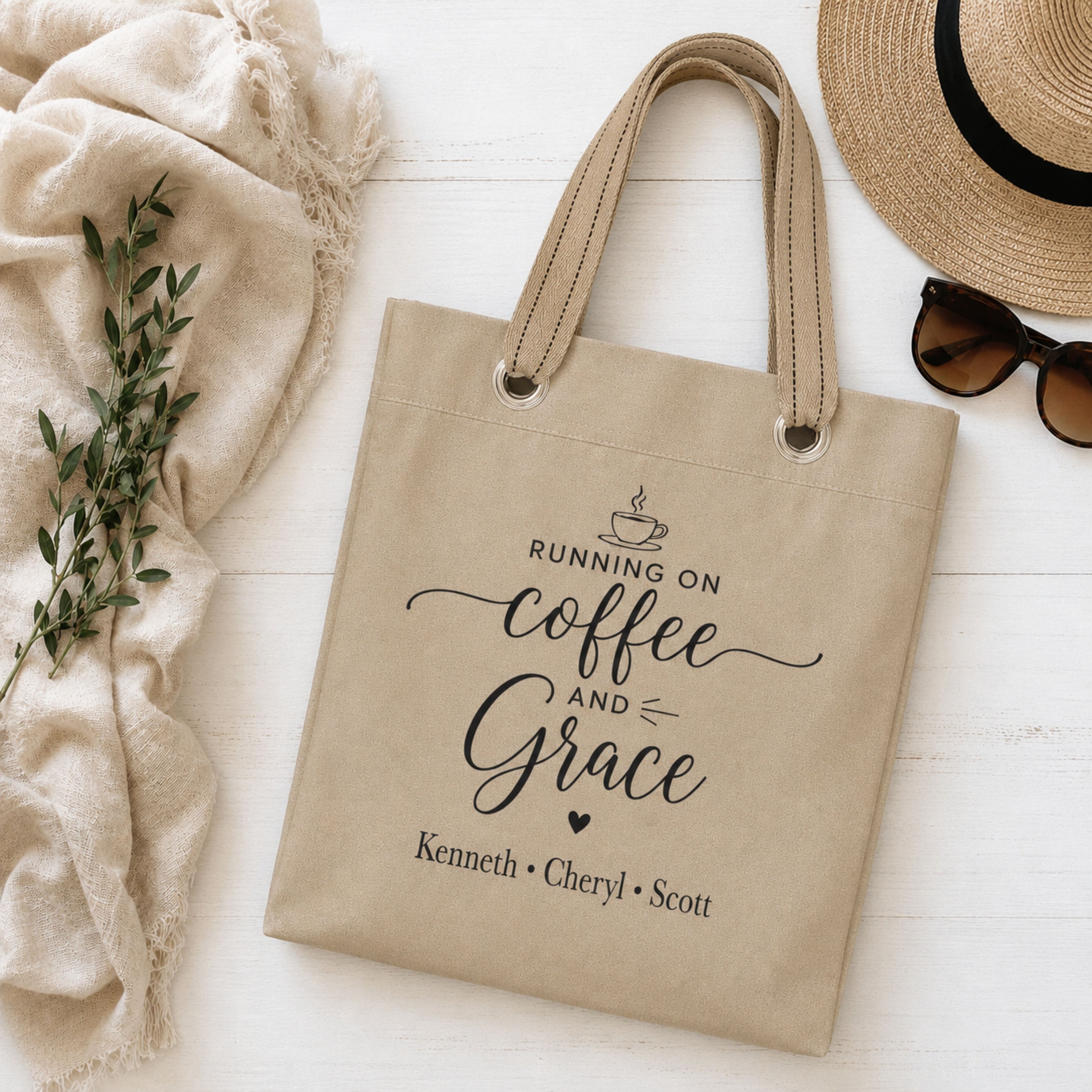 Beige tote bag with 'Running on Coffee and Grace' text and kids names, placed on a light wooden surface with a hat, sunglasses, and plant.
