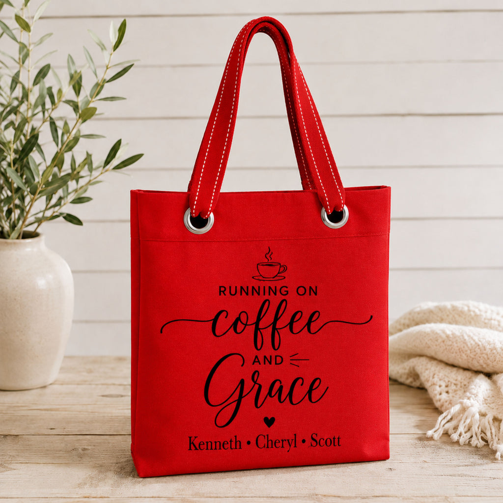 Red tote bag with 'Running on Coffee and Grace' text and kids names on a wooden surface with a plant and blanket.