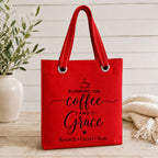 Red tote bag with 'Running on Coffee and Grace' text and kids names on a wooden surface with a plant and blanket.