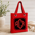 Red tote bag with monogram 'ACR' on a wooden surface with a plant and blanket in the background