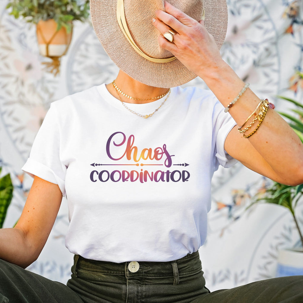 Female sitting down wearing white t-shirt with Chaos Coordinator written in multicolored letters in center of tee