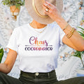 Female sitting down wearing white t-shirt with Chaos Coordinator written in multicolored letters in center of tee