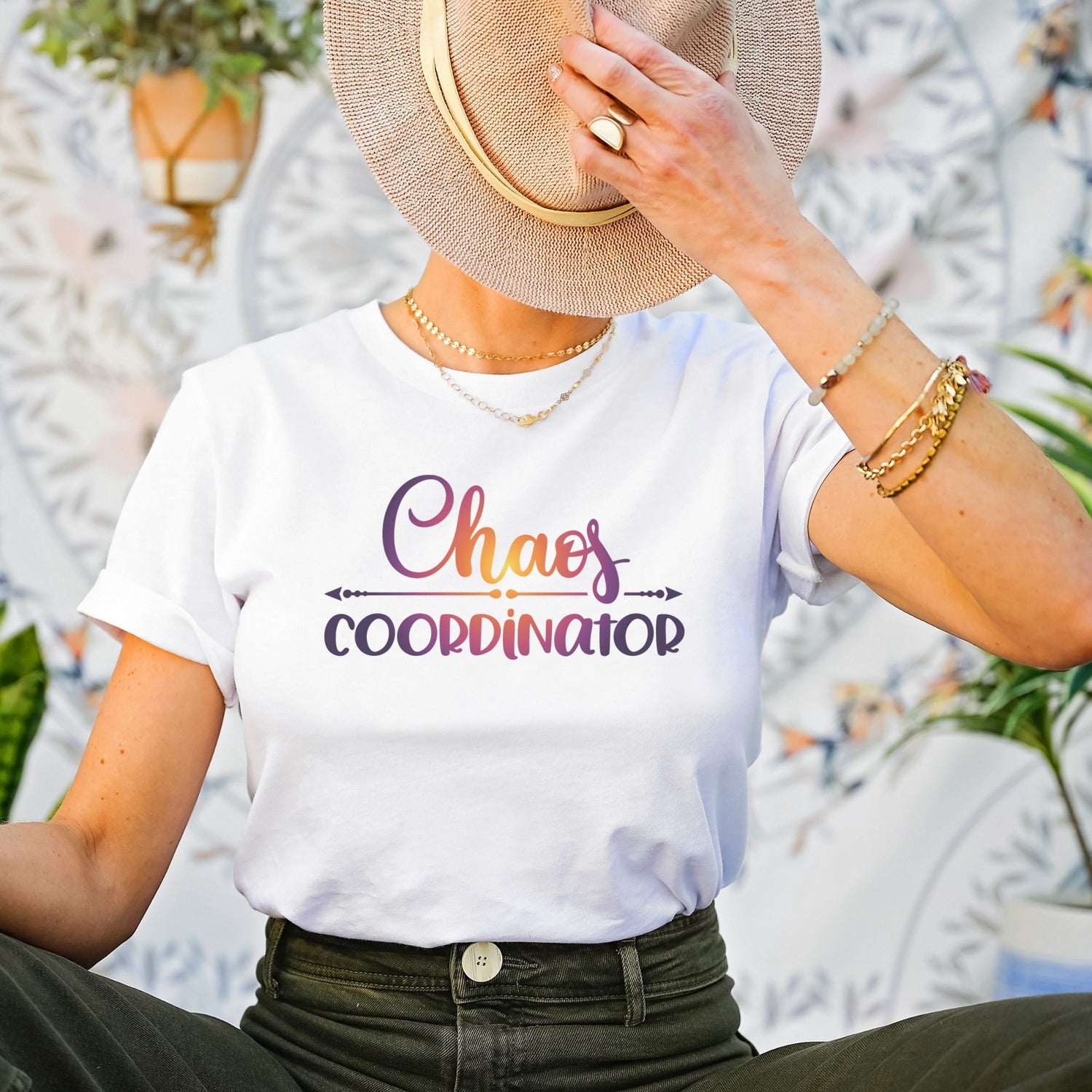 Female sitting down wearing white t-shirt with Chaos Coordinator written in multicolored letters in center of tee