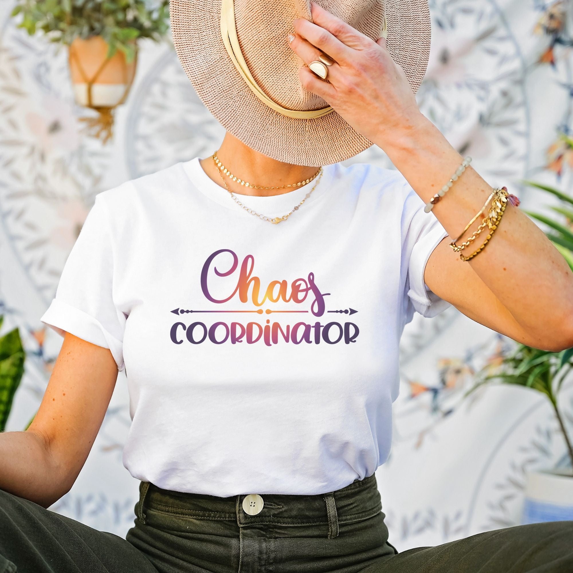 Female sitting down wearing white t-shirt with Chaos Coordinator written in multicolored letters in center of tee